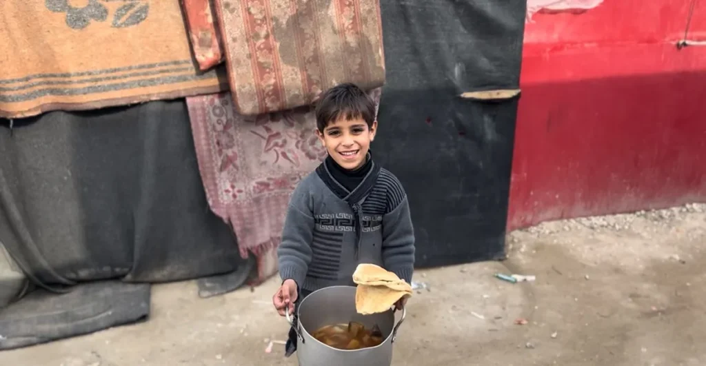 Jonge jongen in Gaza glimlacht terwijl hij een pan met voedsel vasthoudt in een vluchtelingensetting