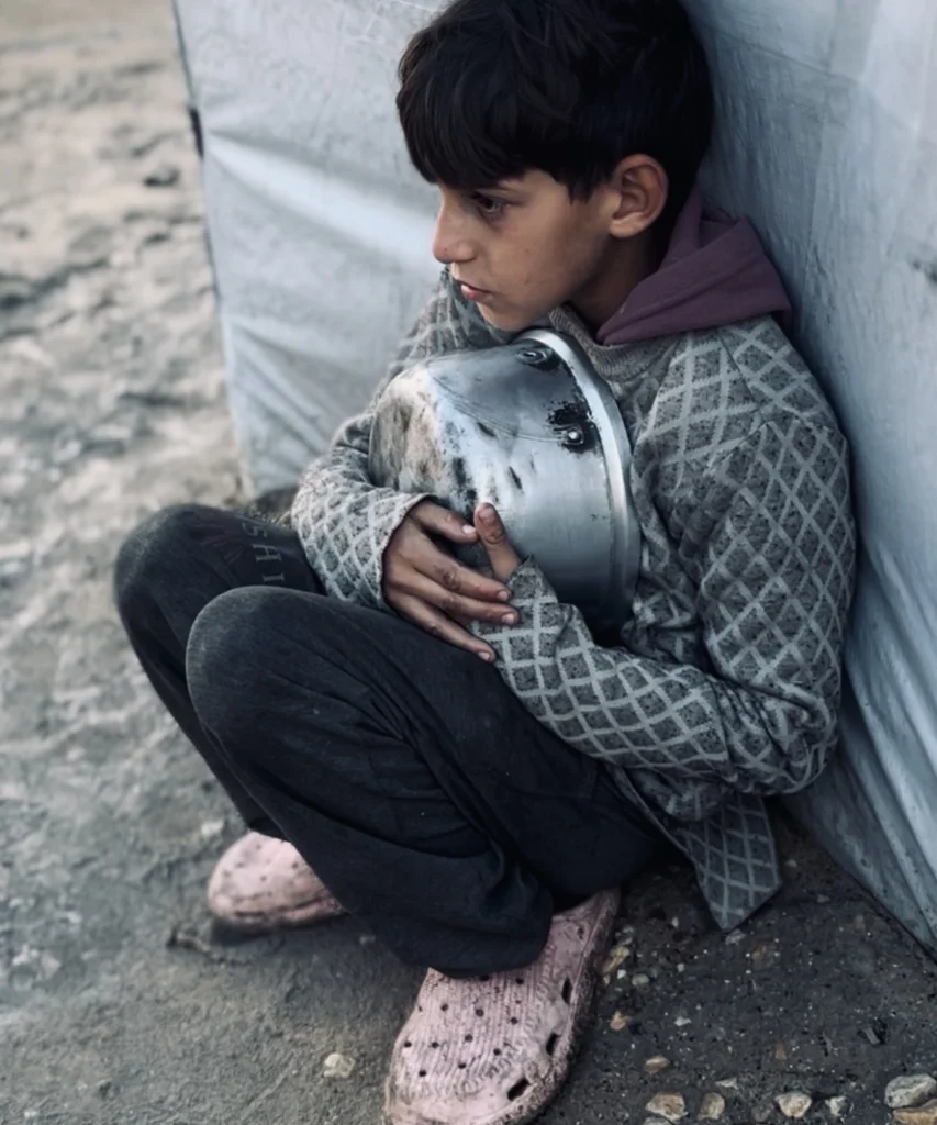 Jongen in een vluchtelingenkamp zit op de grond en houdt een lege, vuile kookpan tegen zijn borst