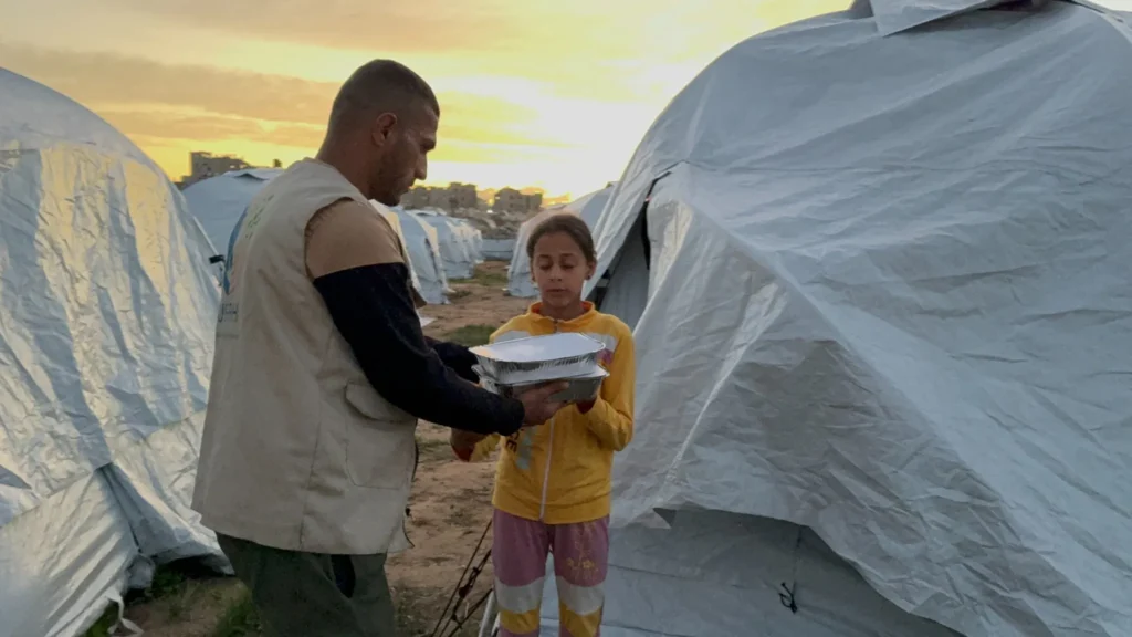 Een hulpverlener overhandigt een warme maaltijd aan een meisje in een vluchtelingenkamp bij zonsondergang, tussen rijen tenten.