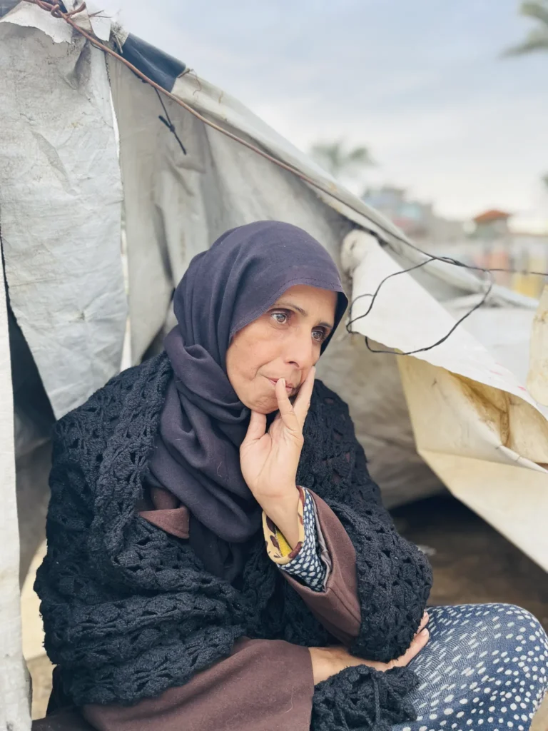 Vrouw in donkere kleding zit voor een geïmproviseerde tent in Gaza, kijkt bezorgd in de verte