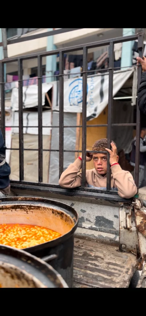 Jongen achter hek kijkt verlangend naar pan met warme maaltijd tijdens voedseluitdeling in Palestina.