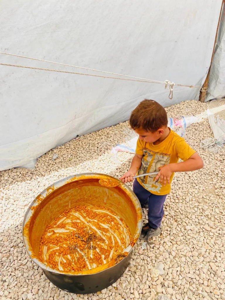 Jonge Palestijnse jongen in vluchtelingenkamp roert in grote pan met restanten van maaltijd