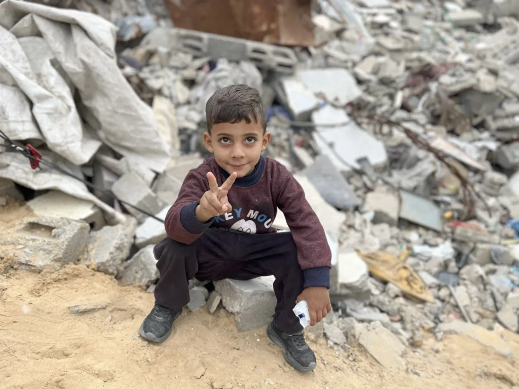 Jongen zit op puin in Gaza en maakt vredesteken met zijn hand, voor ingestorte gebouwen op de achtergrond