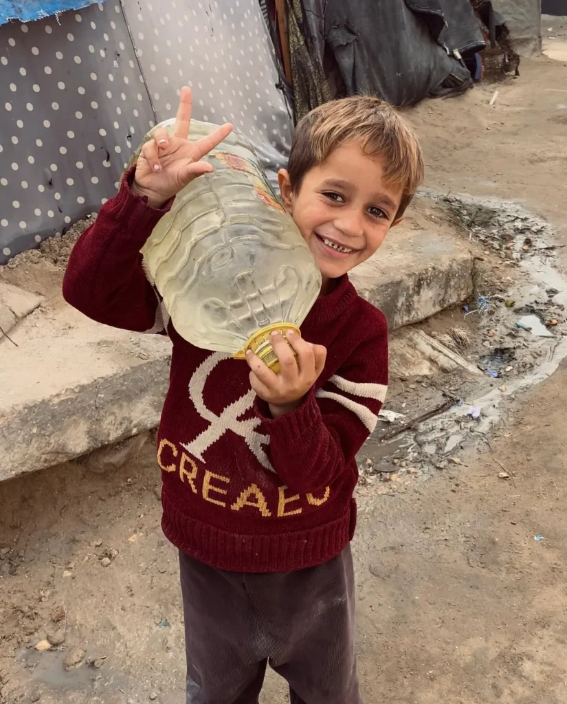 Jongen met een grote fles water glimlacht en maakt een vredesteken in vluchtelingenkamp