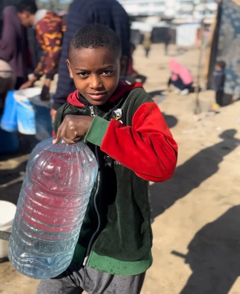 Jonge jongen in vluchtelingengebied tilt een grote jerrycan met water terwijl anderen op de achtergrond wachten