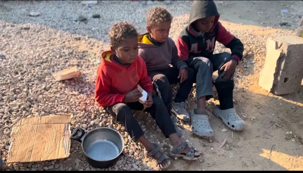 Drie jonge kinderen zitten op de grond met lege pannen en karton bij zich, wachtend op voedsel in een kampomgeving