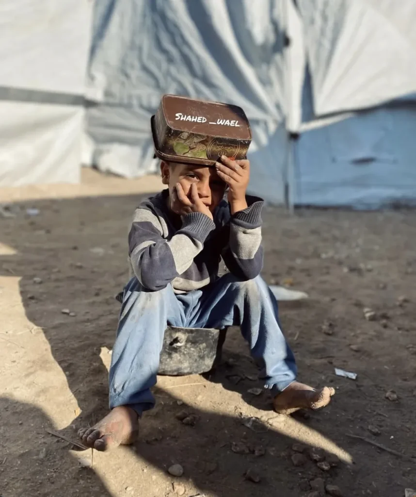 Jongen zit op de grond in een kamp en houdt een blik op zijn hoofd
