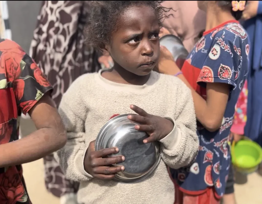 Kind in versleten kleding houdt een lege metalen kom vast in een rij met andere kinderen in een vluchtelingengebied