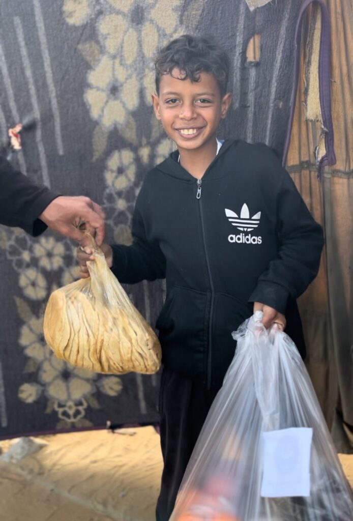 Jonge jongen ontvangt voedselhulp in een tentenkamp, lachend met plastic zakken in zijn handen