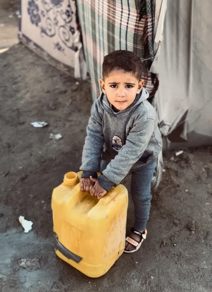 Jonge jongen in grijze hoodie tilt een grote gele jerrycan met water voor een tent.