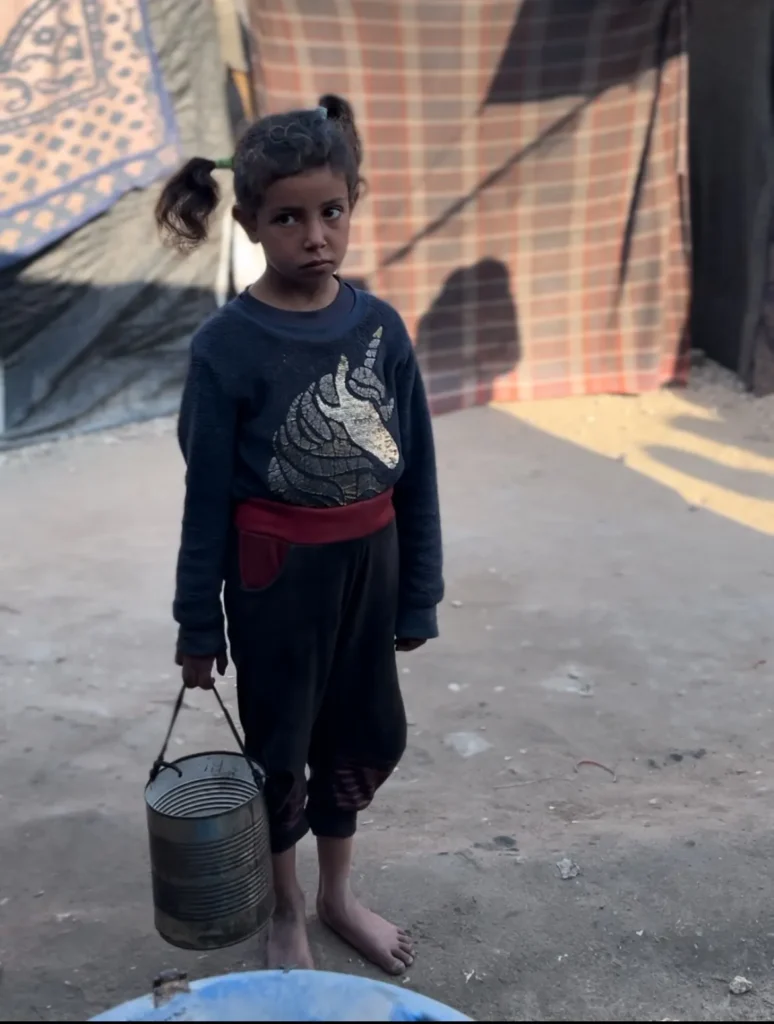 Jong meisje op blote voeten met lege blik, draagt metalen emmer in een geïmproviseerde buitenruimte.