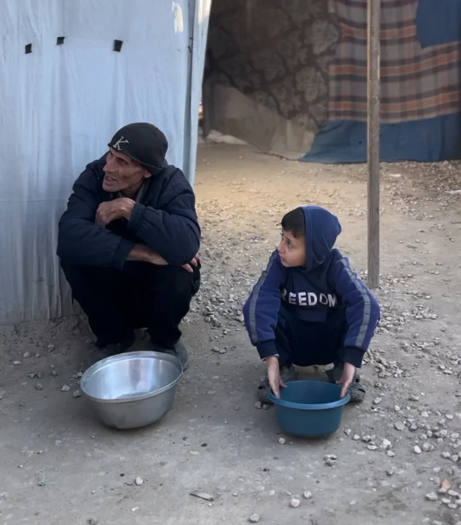 Man en kind hurken met lege kommen bij een tent in een kamp.