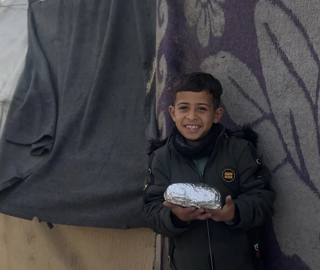 Smiling young boy holding a foil-wrapped meal in front of a tent wall.