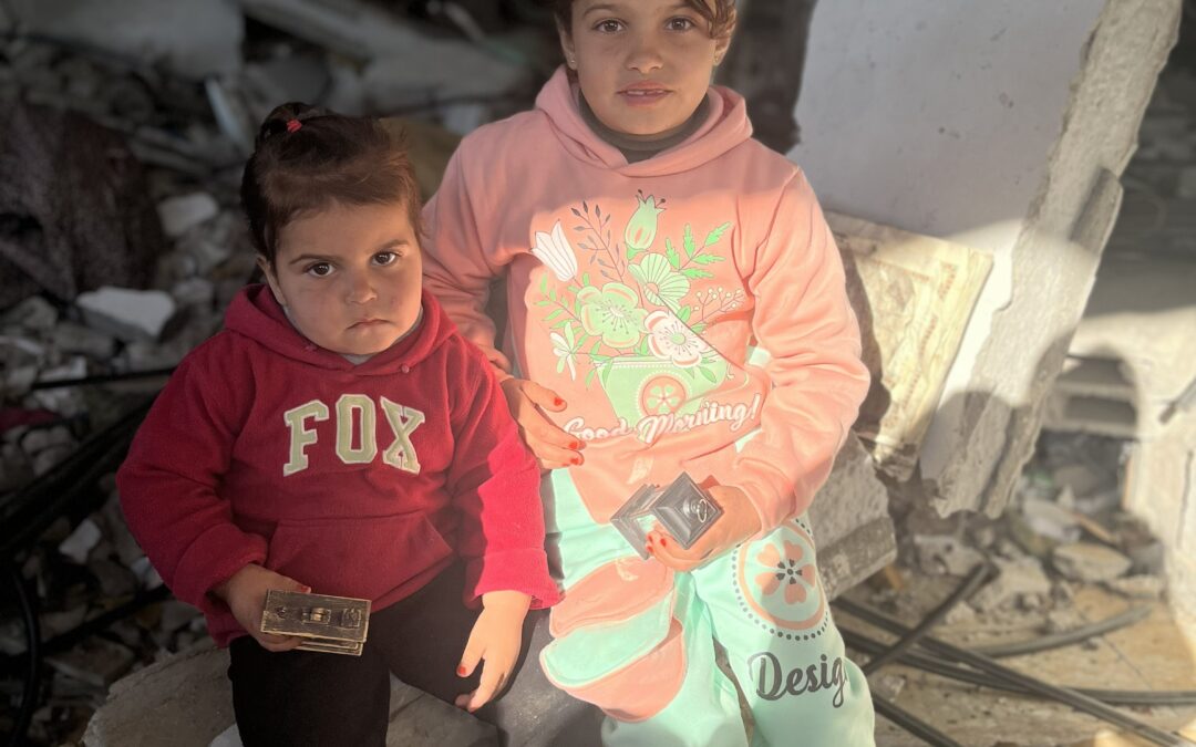 Twee jonge kinderen zitten op puin in een verwoeste omgeving met zonlicht op hun gezicht en houden kleine voorwerpen in hun handen.