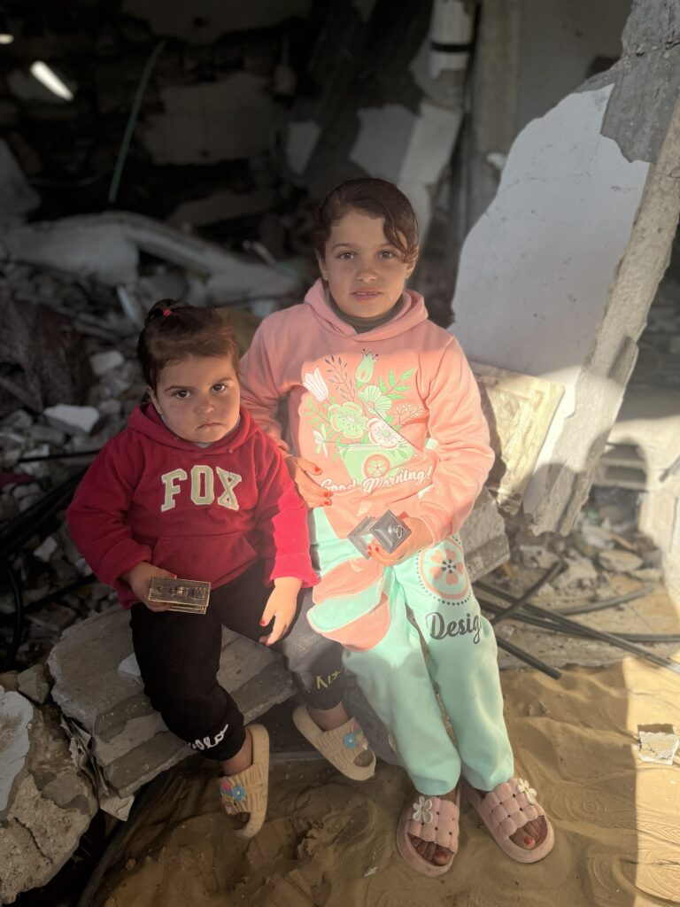 Twee jonge kinderen zitten op puin in een verwoeste omgeving met zonlicht op hun gezicht en houden kleine voorwerpen in hun handen.