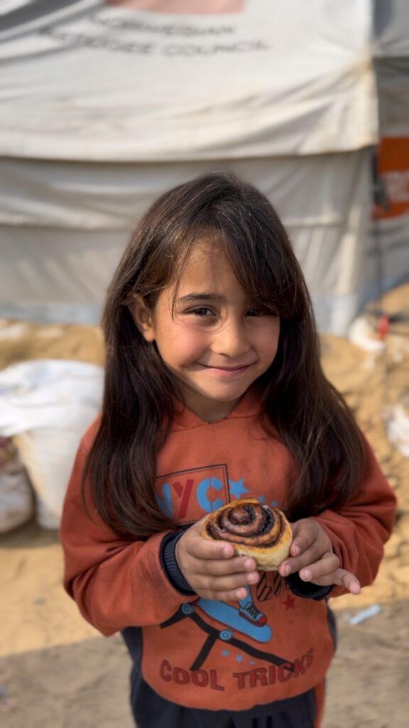Meisje in oranje trui houdt een vers kaneelbroodje vast tijdens voedseluitdeling van Stichting ULFAH; haar glimlach toont de impact van hulp.