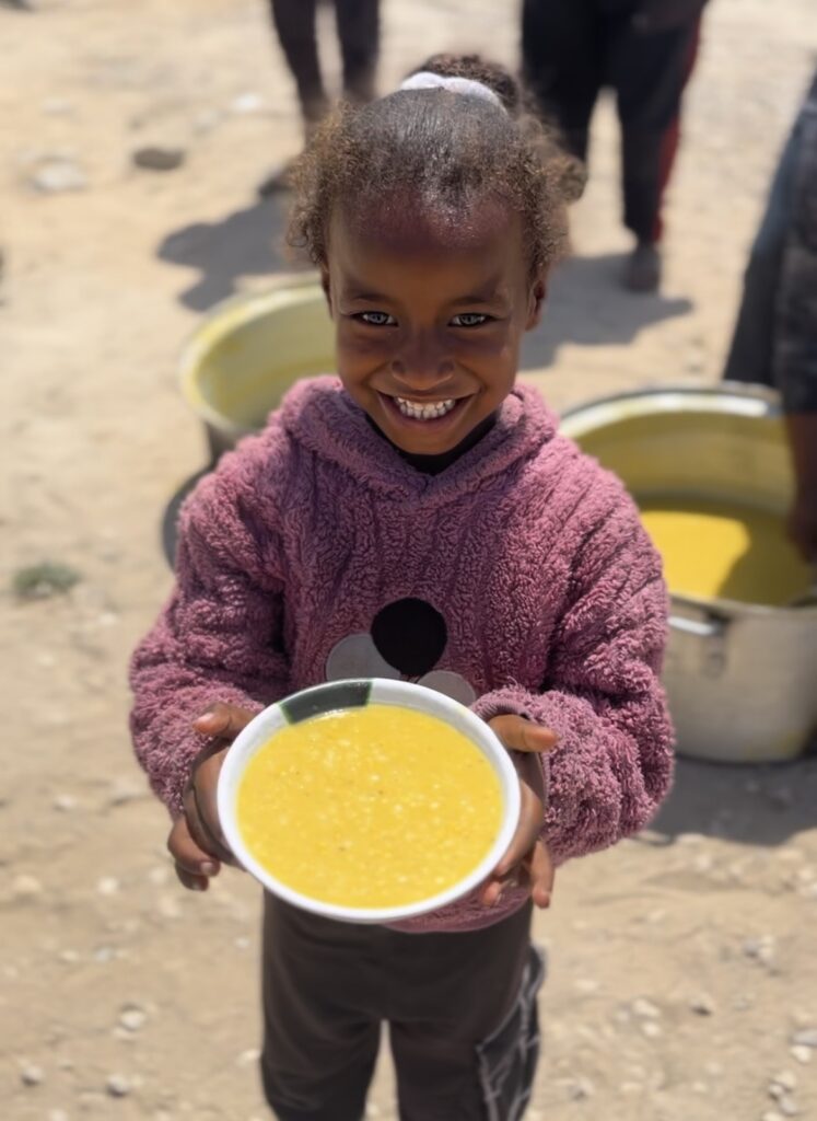 Lachend kind houdt een kom met warme soep vast tijdens een voedselactie van Stichting ULFAH, symbool voor hoop en hulp.
