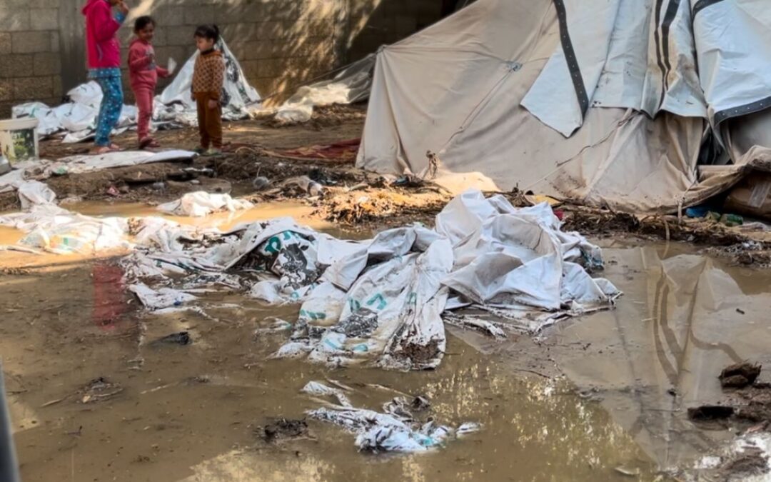 Drie kinderen naast een noodtent, omringd door modderplassen en natte zeilen in een geïmproviseerd kamp